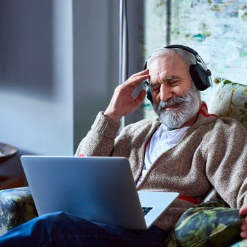 Senior man wearing headphones and using a laptop reclines in armchair as he streams a movie at home