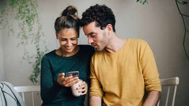 Smiling girlfriend showing smart phone to boyfriend while having coffee in living room