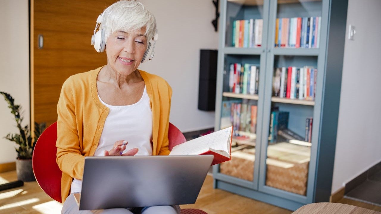 An older woman wearing headphones and holding a book talks before a laptop computer.