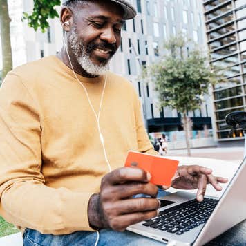 Smiling man sites on a park bench holding a credit card while looking at his laptop.
