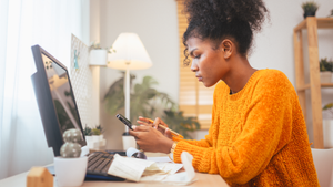 A woman in a yellow sweater is focused on her smartphone, with a computer monitor and notepad on the desk in a bright room.