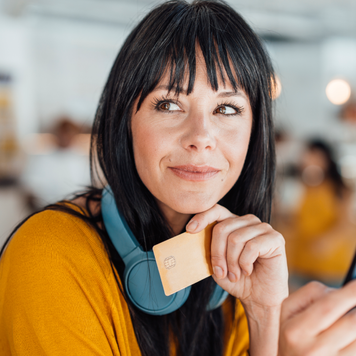 Contemplative woman with with headphones around neck holding credit card and smart phone - stock photo